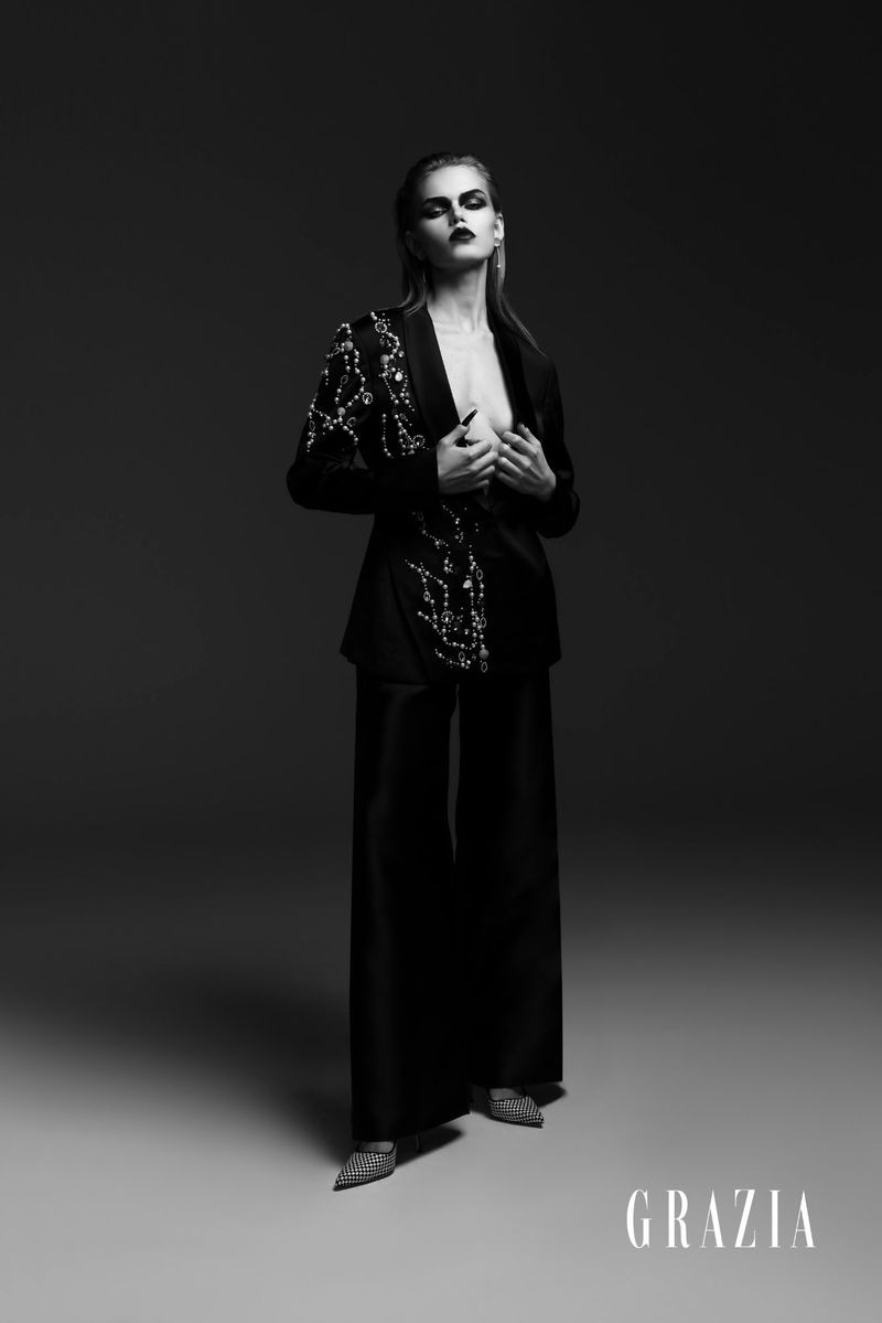 An adult female fashion model poses in a black jacket with decorative embellishments and wide-leg pants in a photo studio setting.