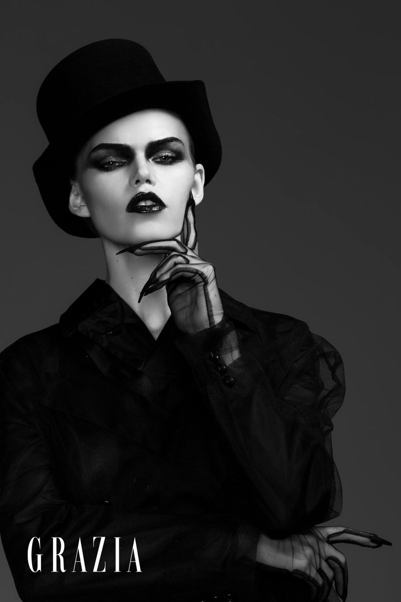 Fashion portrait of a woman with dramatic eye makeup and dark lipstick wearing a black top hat, mesh gloves, and sheer jacket.