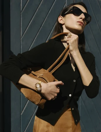 Woman in black cardigan and brown skirt with suede purse, ornamental wrist-watch, sunglasses, and rings poses against blue door.