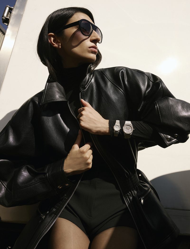Model wearing a black leather jacket and sunglasses with two silver wristwatches against a white background