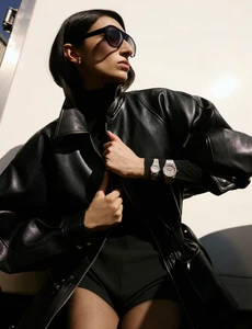 Model wearing a black leather jacket and sunglasses with two silver wristwatches against a white background