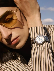 Close-up of a model wearing yellow-tinted sunglasses and a striped shirt, showcasing a silver wristwatch on her arm against a clear sky