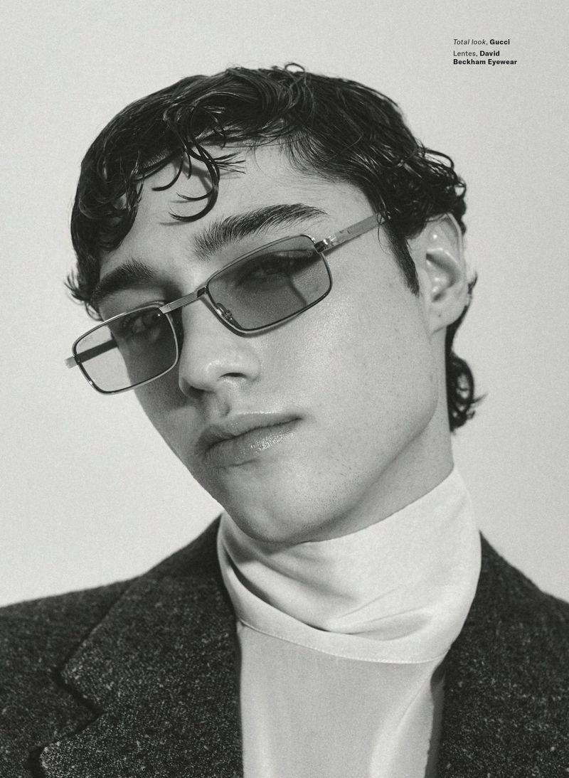 Monochrome portrait of a young adult man with dark hair, wearing glasses, a turtleneck sweater, and a dark jacket, against a white background.