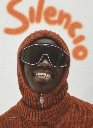Headshot of a man with dark skin wearing futuristic sunglasses and a rust-colored turtleneck with the word 'Silencio' spray-painted behind him.