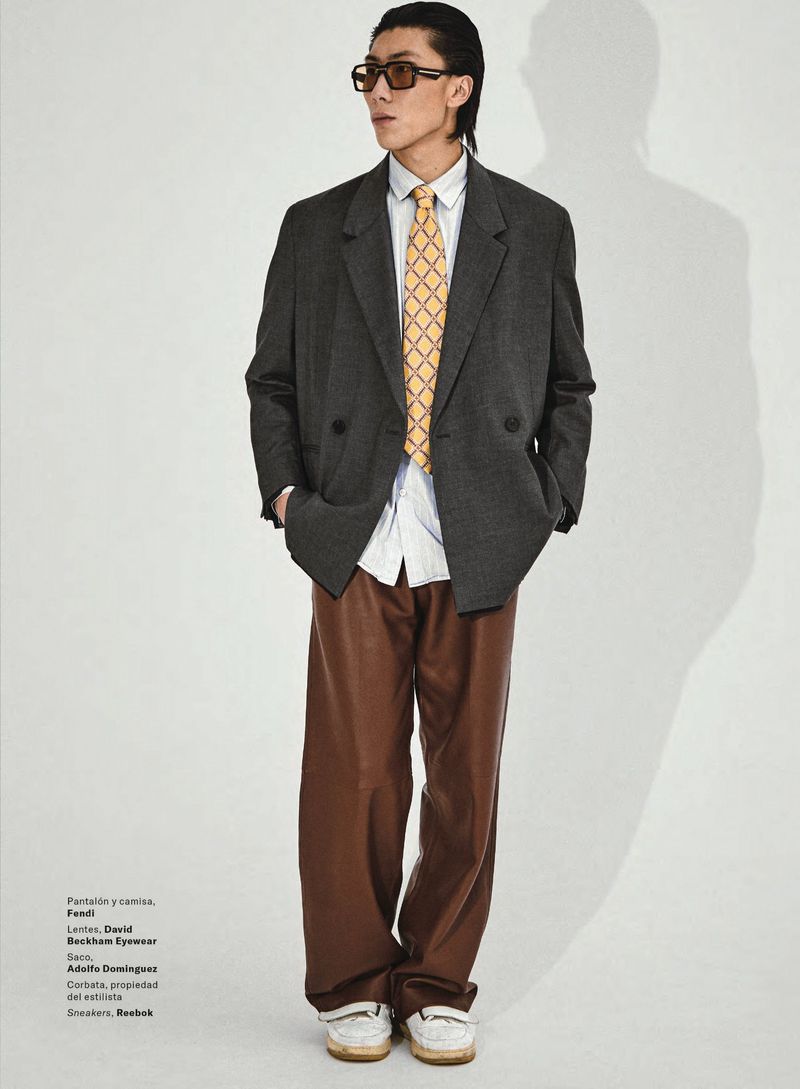 A full-length portrait of a young adult male in a gray blazer, white collared shirt, brown leather pants, and white sneakers, on a white background.