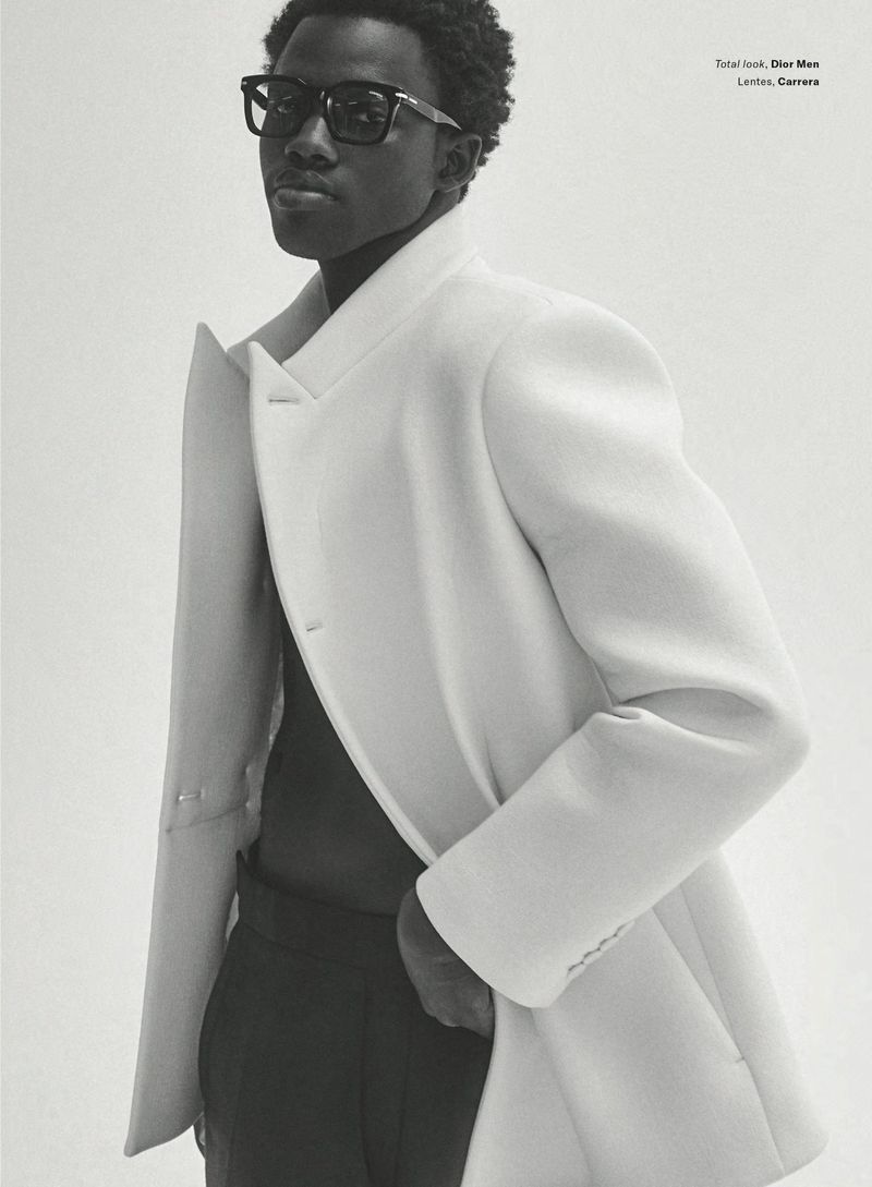 Man wearing a structured white jacket and thick black sunglasses against a white background