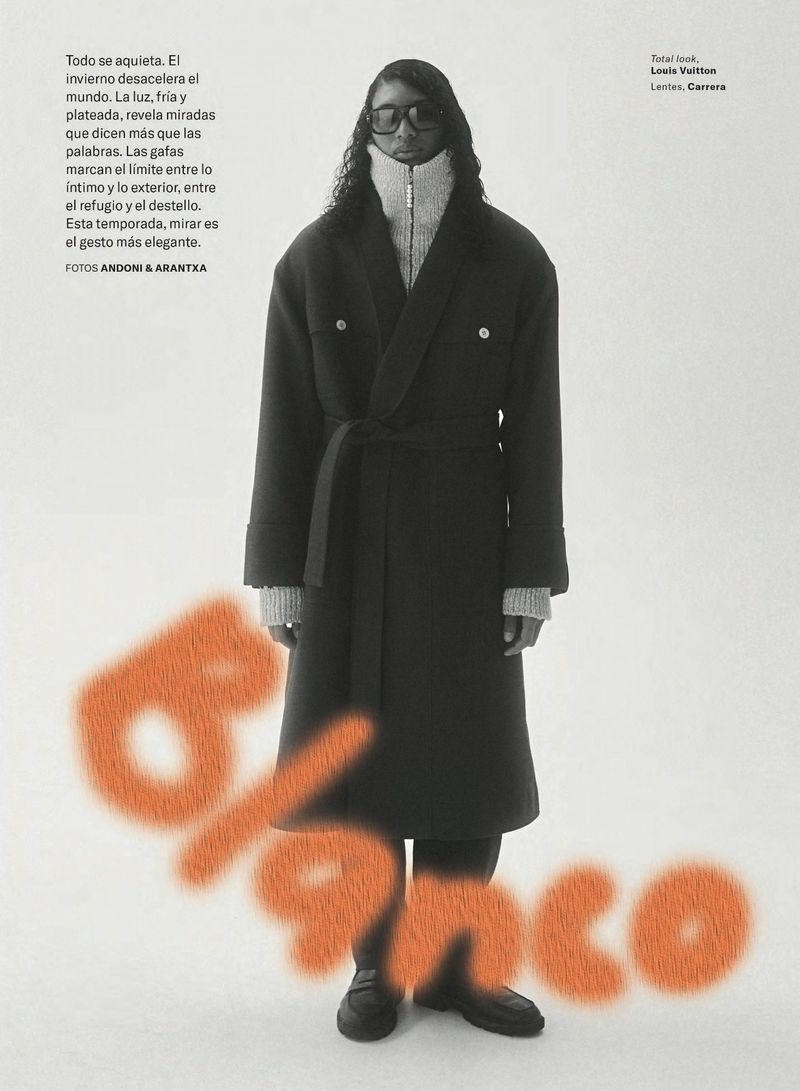 A person poses in an elegant dark coat, light turtleneck sweater, and black shoes against a bright background with a spray-painted word.
