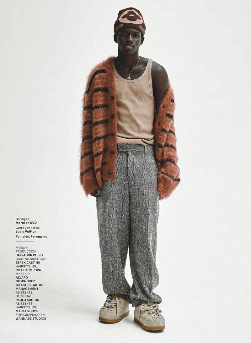 Male model wears a brown striped cardigan, tan tank top, gray tweed trousers, and sneakers in a full-length fashion studio shot.