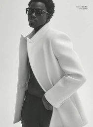 Man wearing a structured white jacket and thick black sunglasses against a white background