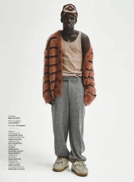 Black man wearing a brown beanie, fuzzy striped cardigan, beige tank top, grey trousers, and platform sneakers on white background.