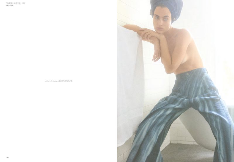 Adult female poses in wide-leg pants and a towel on her head near a bathtub with white-toned, diffused studio lighting.