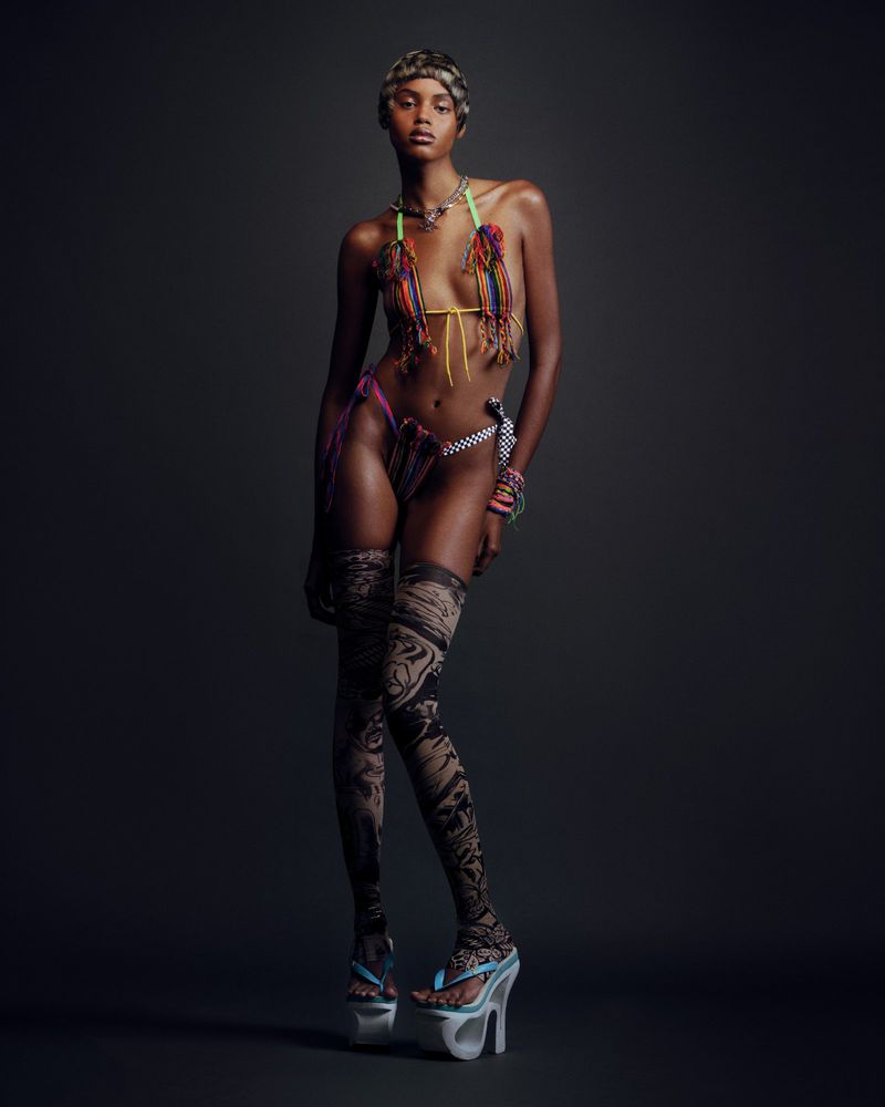 Fashion shot of an African-descent woman in a decorative bikini, patterned tights, and platform sandals against a solid dark background.