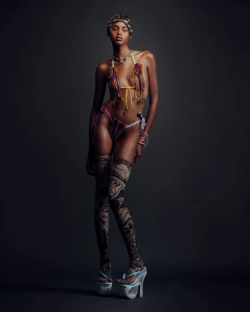 Fashion shot of an African-descent woman in a decorative bikini, patterned tights, and platform sandals against a solid dark background.