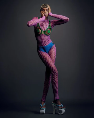 A full-length studio shot of a female model in a pink fishnet bodysuit, posing in front of a grey background, wearing platform flip-flops.