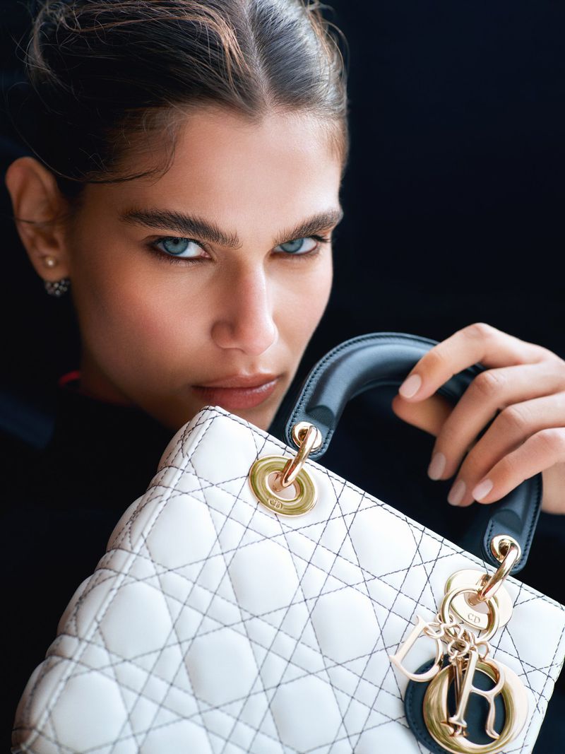 Close-up of a blue-eyed woman with dark hair, posing with a white Lady Dior handbag against a dark background in a fashion editorial.