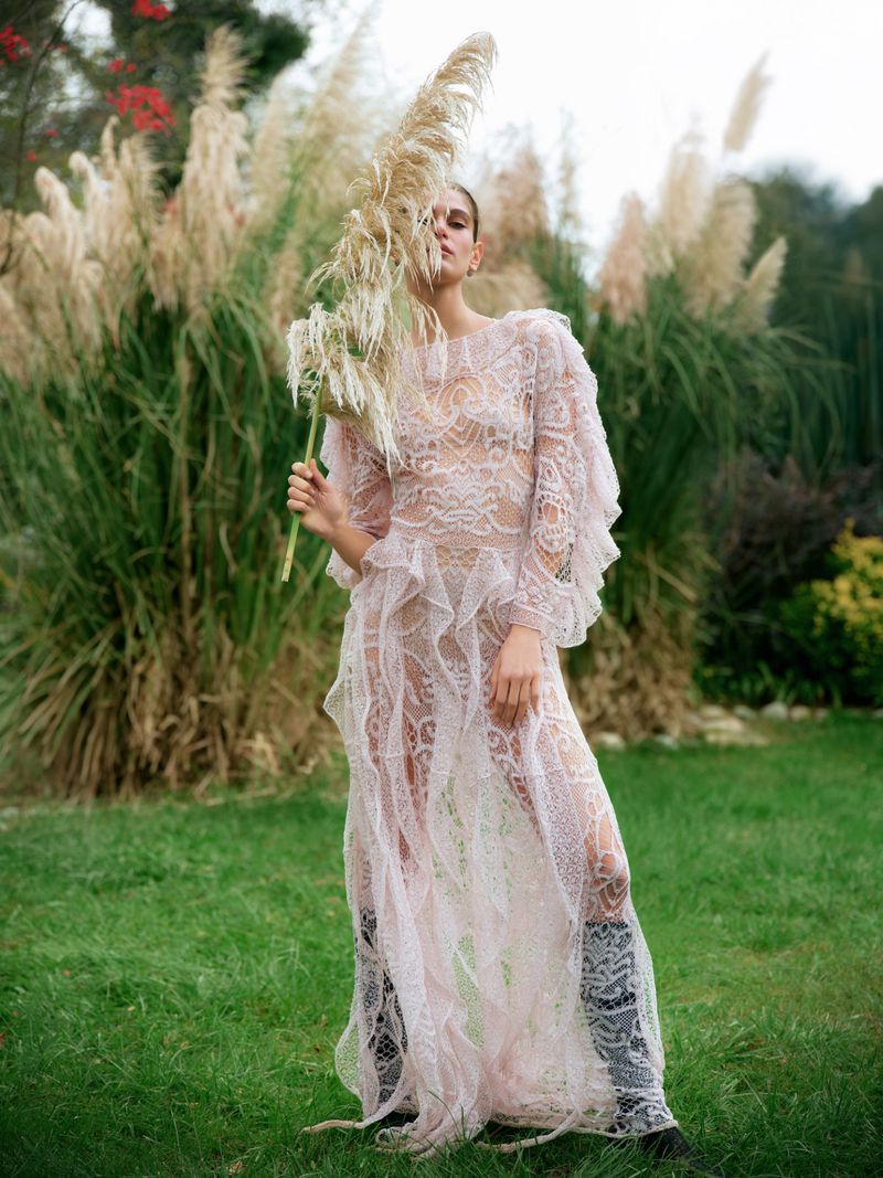 Woman in a lace dress standing in a green field, holding pampas grass in front of her face; the background is overgrown with tall grass.