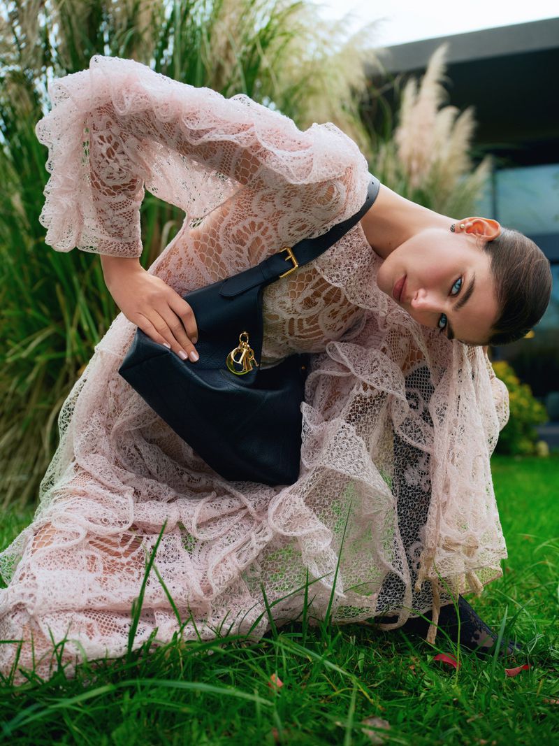 Fashion shot of a woman in a pink lace dress, bending slightly forward, holding a black bag with a gold emblem in a grassy outdoor setting.