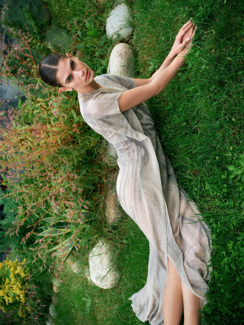 Woman in flowing dress lies on green grass surrounded by lush foliage in an outdoor setting, captured in a top-down view.