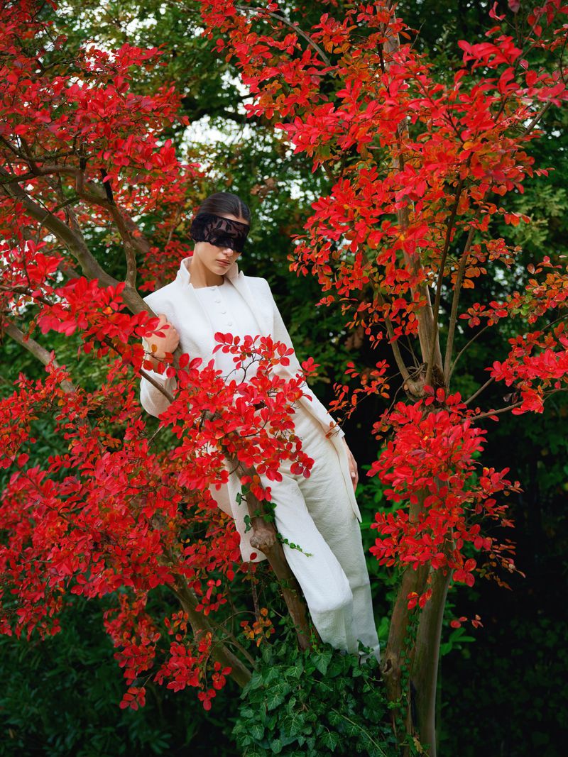 Woman in white suit and lace mask poses in a red-leafed tree against green foliage, creating a whimsical fashion statement in nature.