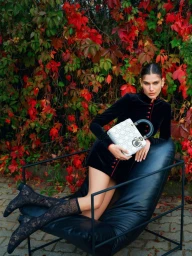 Model wearing a black dress holding a white handbag while lounging on a black leather chair outdoors