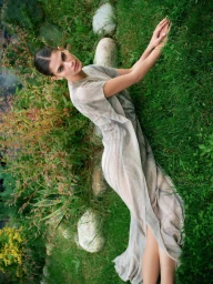 Model wearing a sheer textured gown reclining on a grassy slope with large smooth garden stones.