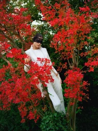 Model in white suit and black lace blindfold posing amidst vibrant red autumn foliage for Elle Turkiye