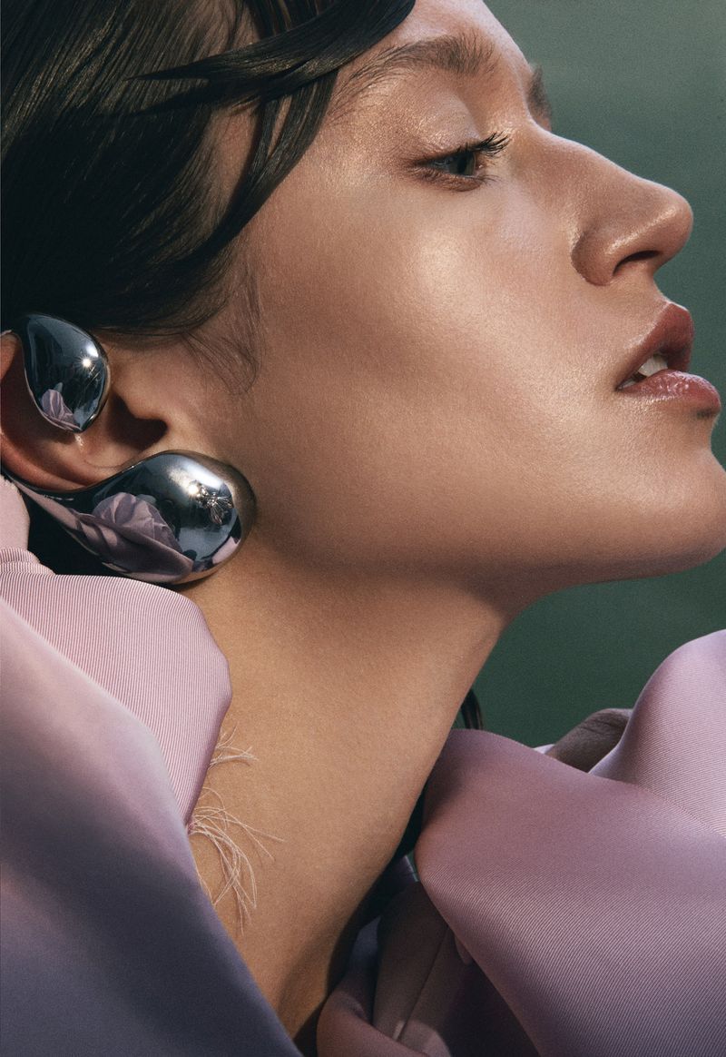 Profile shot of a fashionable adult woman with dark hair wearing large silver teardrop earrings and a light pink blouse against a green backdrop.