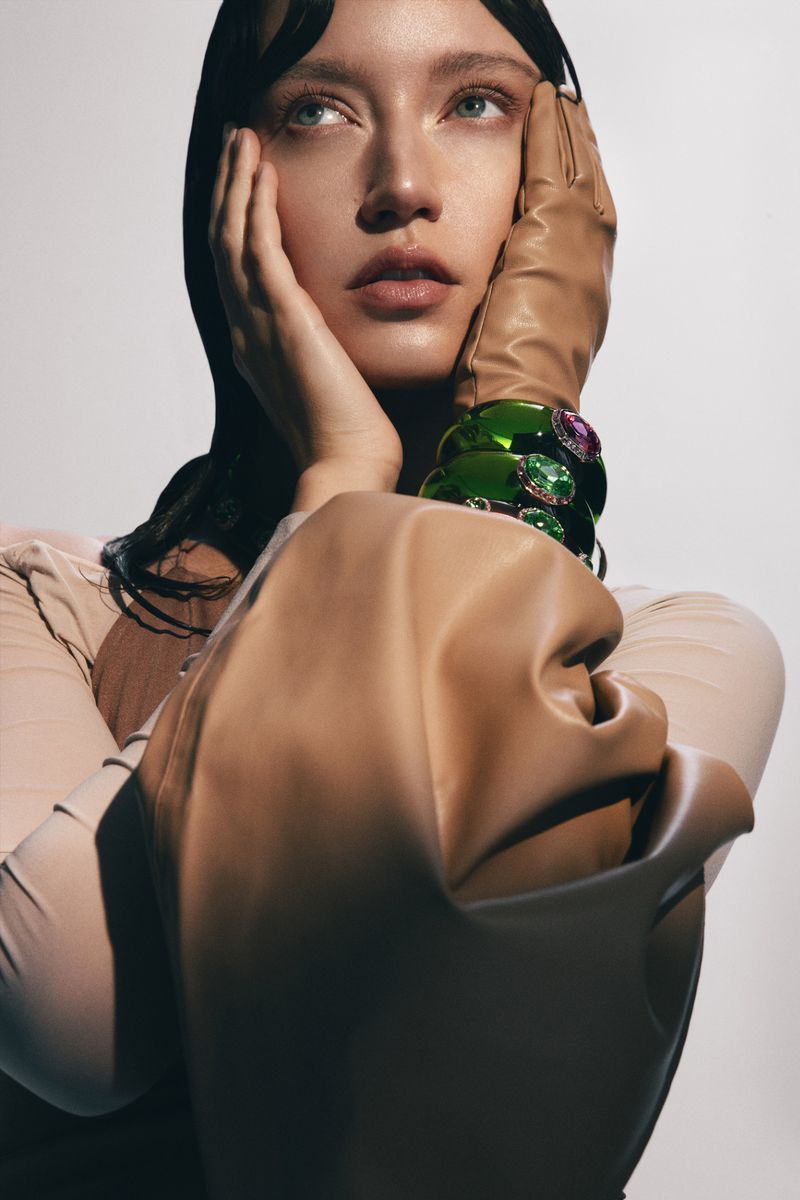 Close-up studio portrait of a woman with hands on her face, wearing leather gloves and bracelets, gazing upward in a well-lit setting.