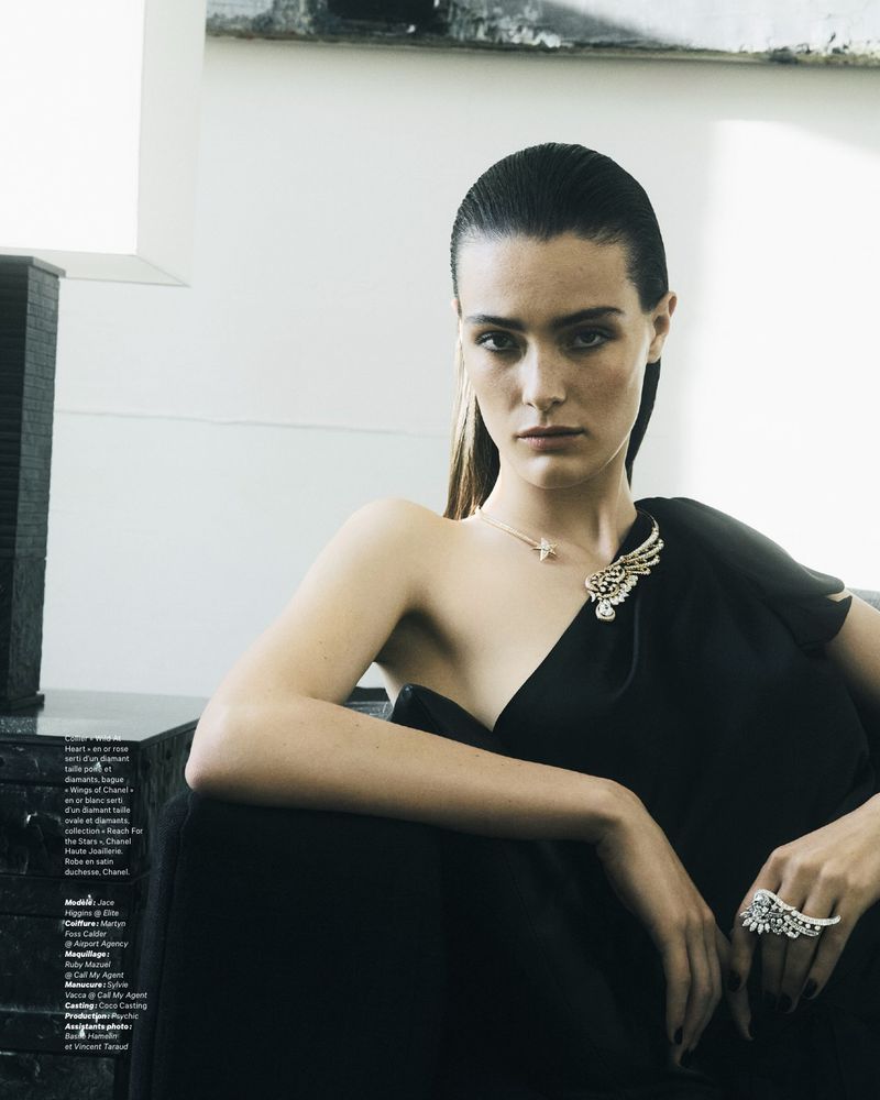 Model with slicked-back dark hair wearing a black dress and diamond jewelry against a light background.