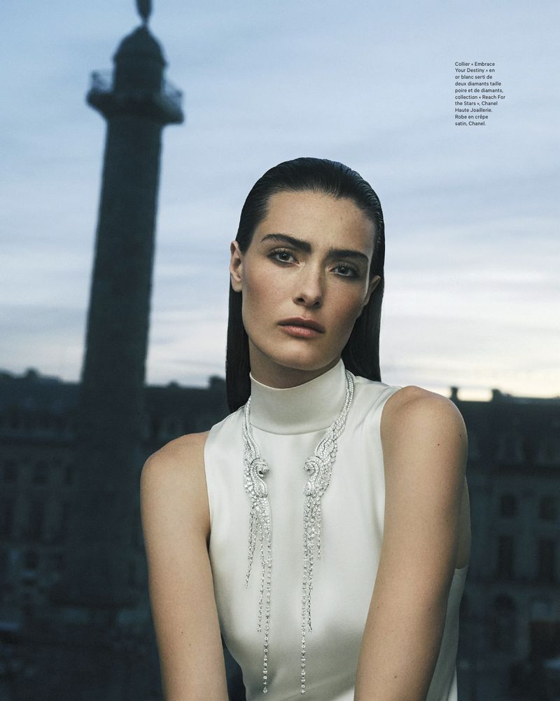 Elegant woman with long dark hair poses in a white dress and diamond necklace, standing against an urban Paris background with a tall monument.