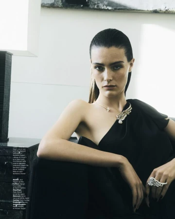 Model with slicked-back dark hair wearing a black dress and diamond jewelry against a light background.