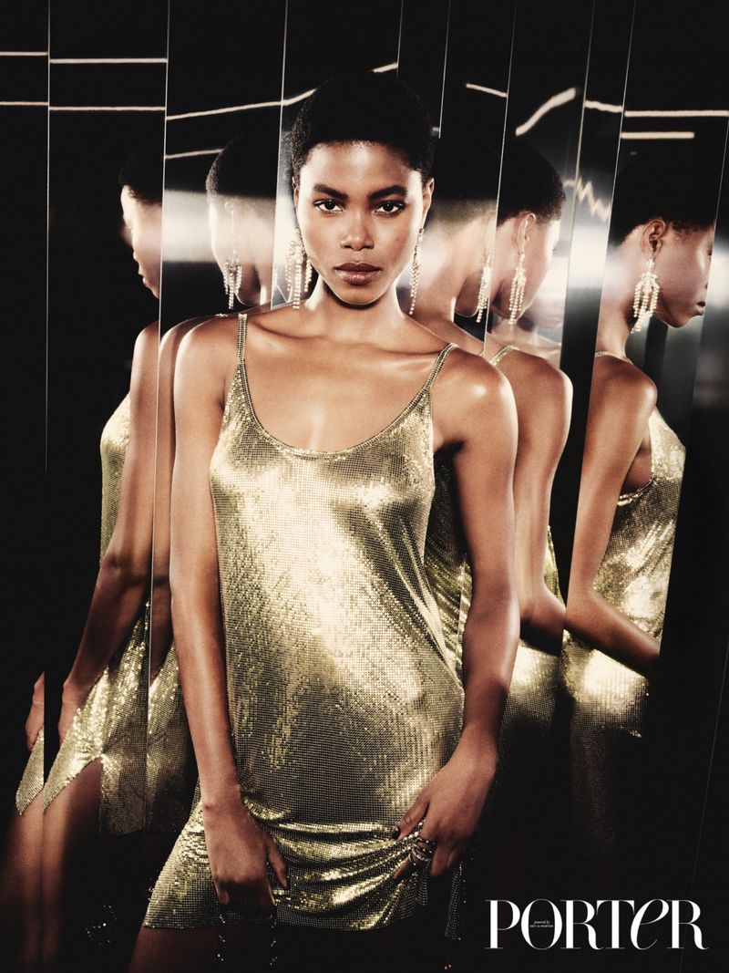 A woman with dark hair poses indoors in a shimmering gold dress. She stands in front of a fractured mirror in a fashion shot.