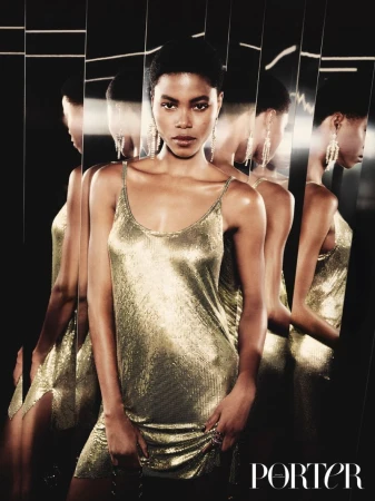 A woman with dark hair poses indoors in a shimmering gold dress. She stands in front of a fractured mirror in a fashion shot.