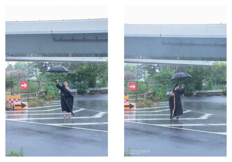 Woman in a black coat holds an umbrella in the rain, standing in a crosswalk under a highway overpass with green foliage.