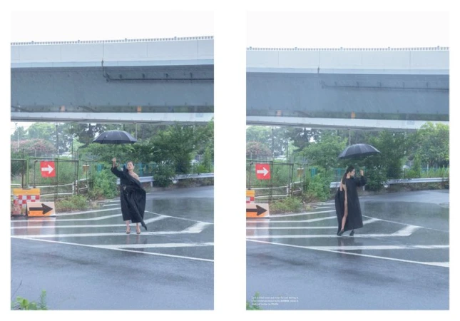Woman in a black coat holds an umbrella in the rain, standing in a crosswalk under a highway overpass with green foliage.
