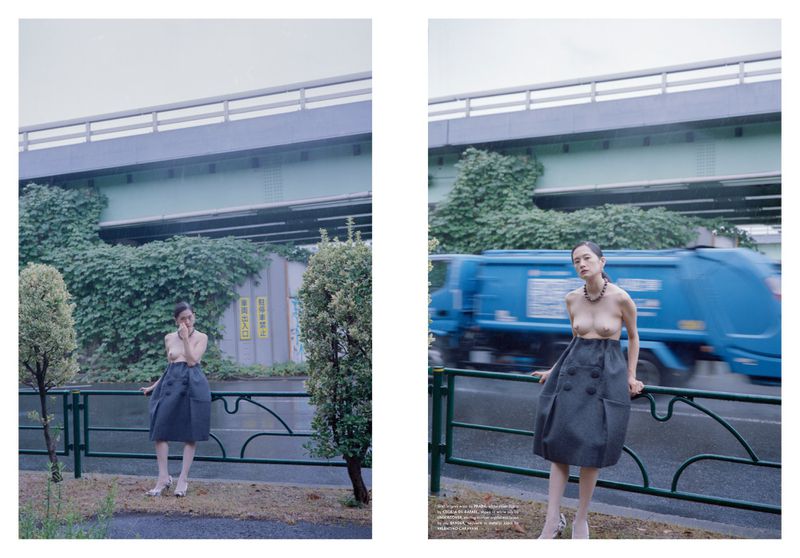 A woman in a grey skirt and beige top stands on a sidewalk in front of a busy street with an overpass and lots of green foliage.