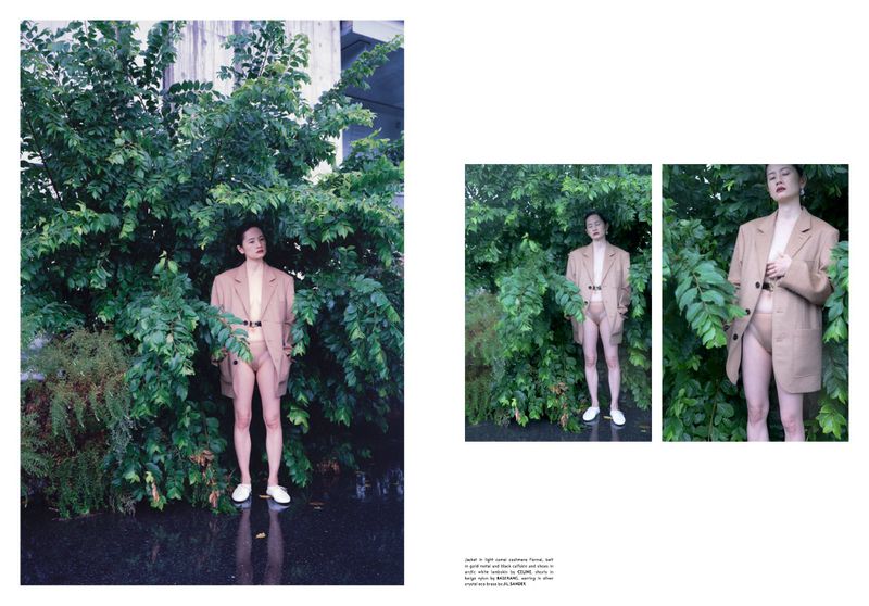 A stylish adult woman stands among lush foliage, wearing a beige blazer, matching bottoms, and white shoes in a fashion editorial shot.