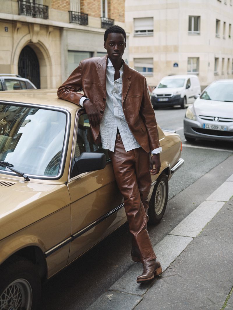 Fashionable man poses with vintage car in city street. He wears a brown leather jacket and pants, a light blue shirt, and brown boots.