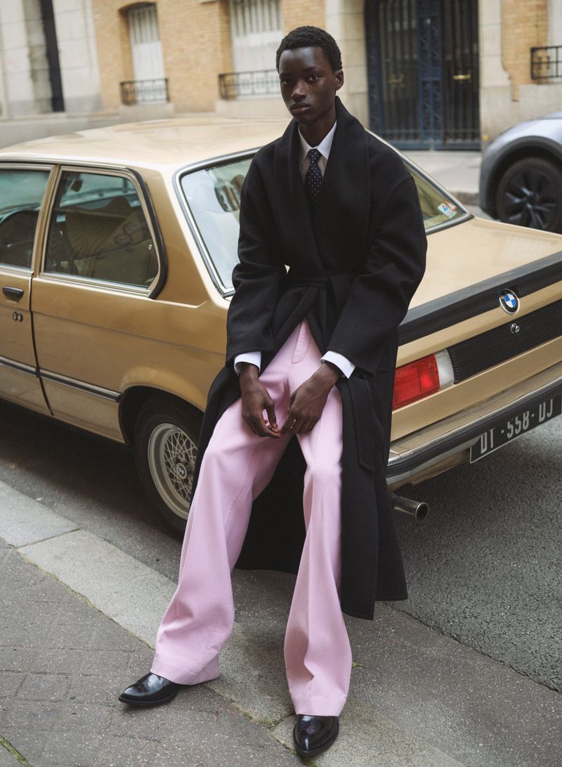 An adult with dark hair and a dark coat leans on a gold car wearing pink pants and black shoes on an urban street.
