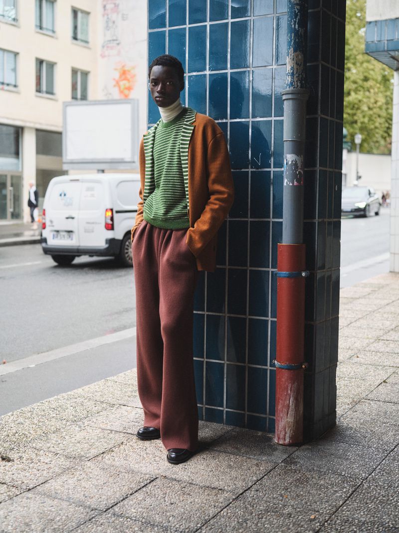 Outdoor fashion shot of an African-American man wearing a cardigan, sweater, and wide-leg trousers, posing against a tiled wall in an urban setting.