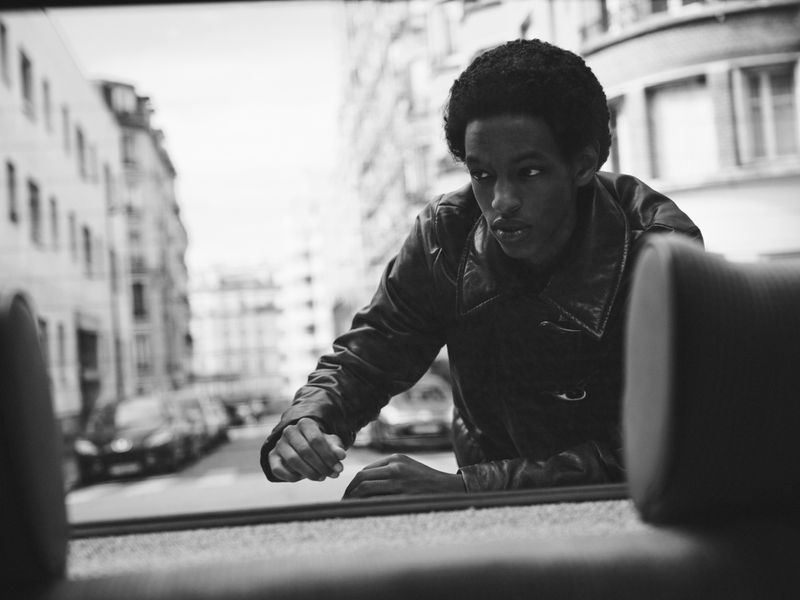 Black and white portrait of a young man in a leather jacket looking out of a car window at a city street with buildings and cars.
