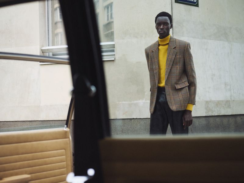 Man in yellow turtleneck and blazer stands outdoors near building and car, showcasing fashion with focus on style and urban environment.