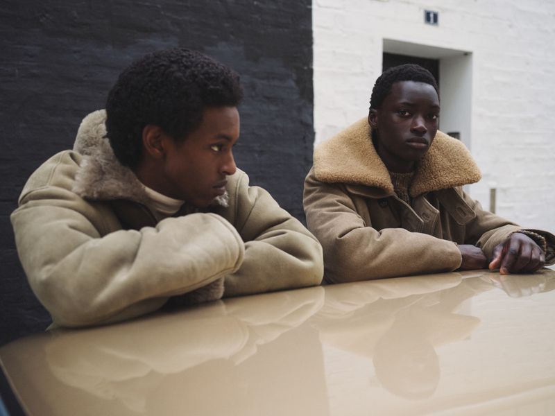 Two young Black men, side-by-side, in shearling-collared coats, pose at a table with neutral background. Neutral, soft lighting, medium shot.