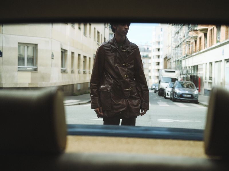 A person in a brown jacket stands in the middle of a city street, seen through the rear window of a car, with buildings visible.