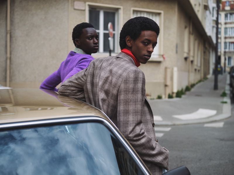 Two Black men in fashionable outfits lean on a vintage car on a European street, showcasing an editorial-style fashion photograph.