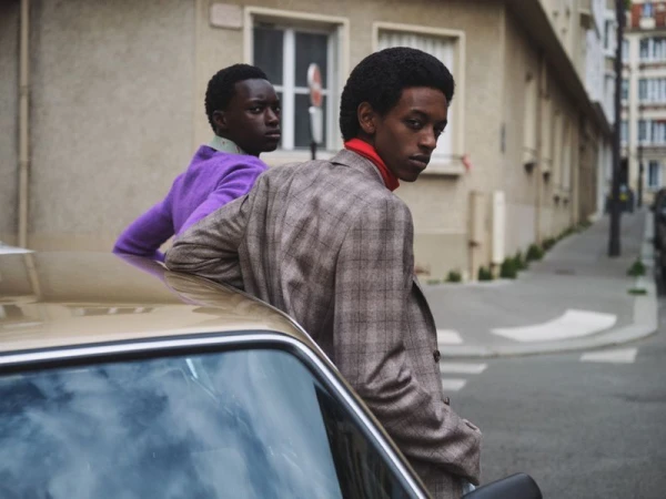 Two Black men in fashionable outfits lean on a vintage car on a European street, showcasing an editorial-style fashion photograph.