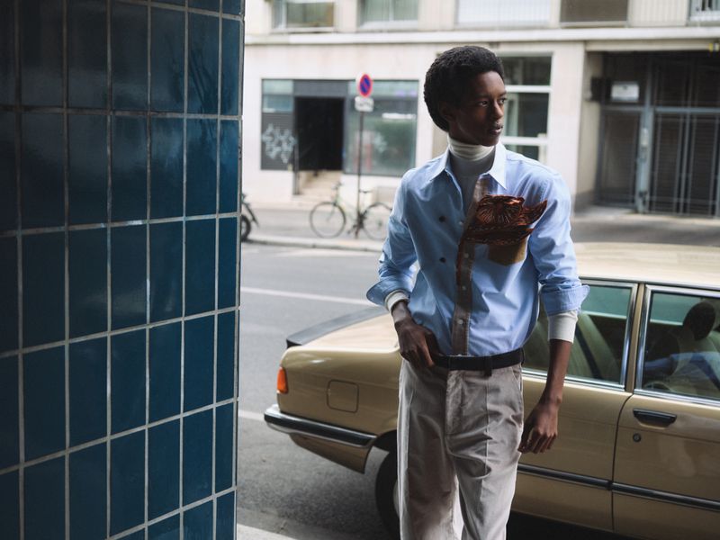 Young man in blue shirt, white turtleneck, and tan pants poses next to a vintage car on a city street, fashion editorial photography.