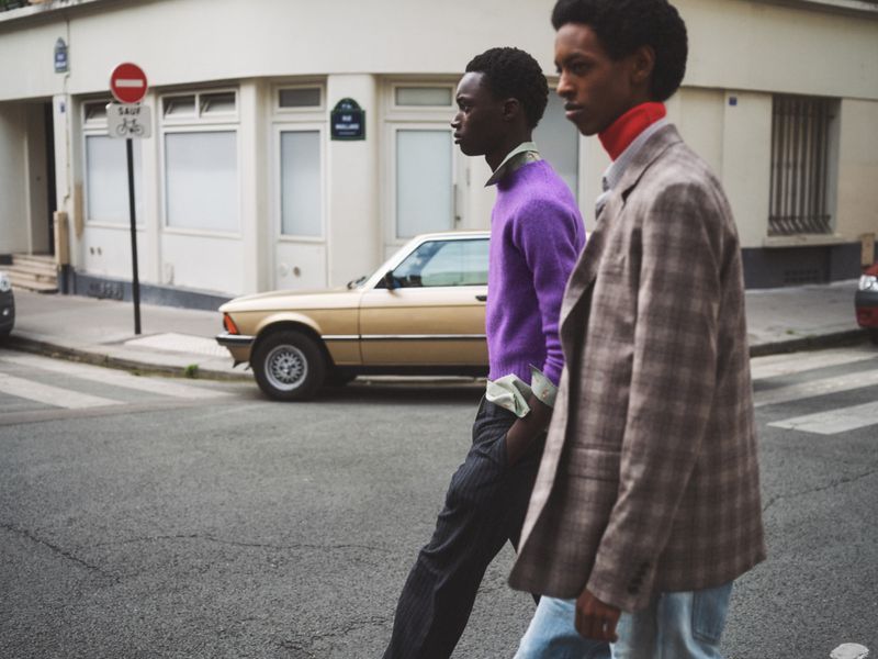 Two Black male fashion models in stylish casual wear walking along an urban crosswalk with vintage car in background.