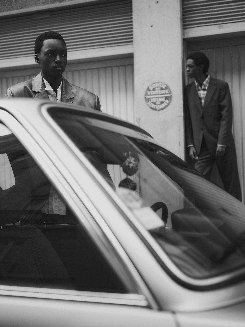 Monochromatic outdoor portrait of two adult Black men in jackets standing near a vintage car on a city street with neutral-toned buildings.