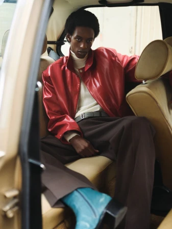 Fashionable man in a red jacket and brown pants sits inside a vintage car with tan interior and turquoise boots, looking stylish.